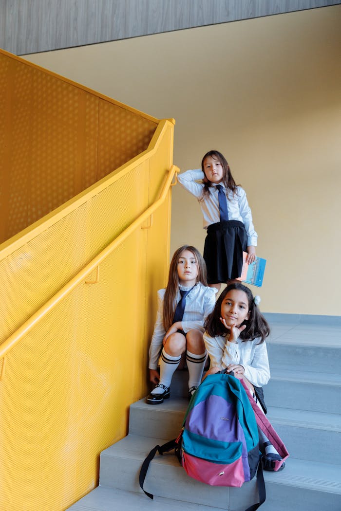 Three young girls in uniforms sitting on a staircase inside a school building.